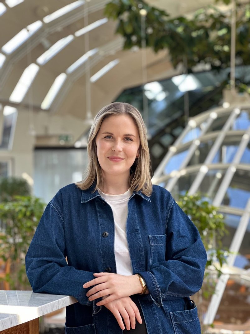 Woman with dark blonde hair, denim jacket and white t-shirt. The woman is leaning against a marble table.